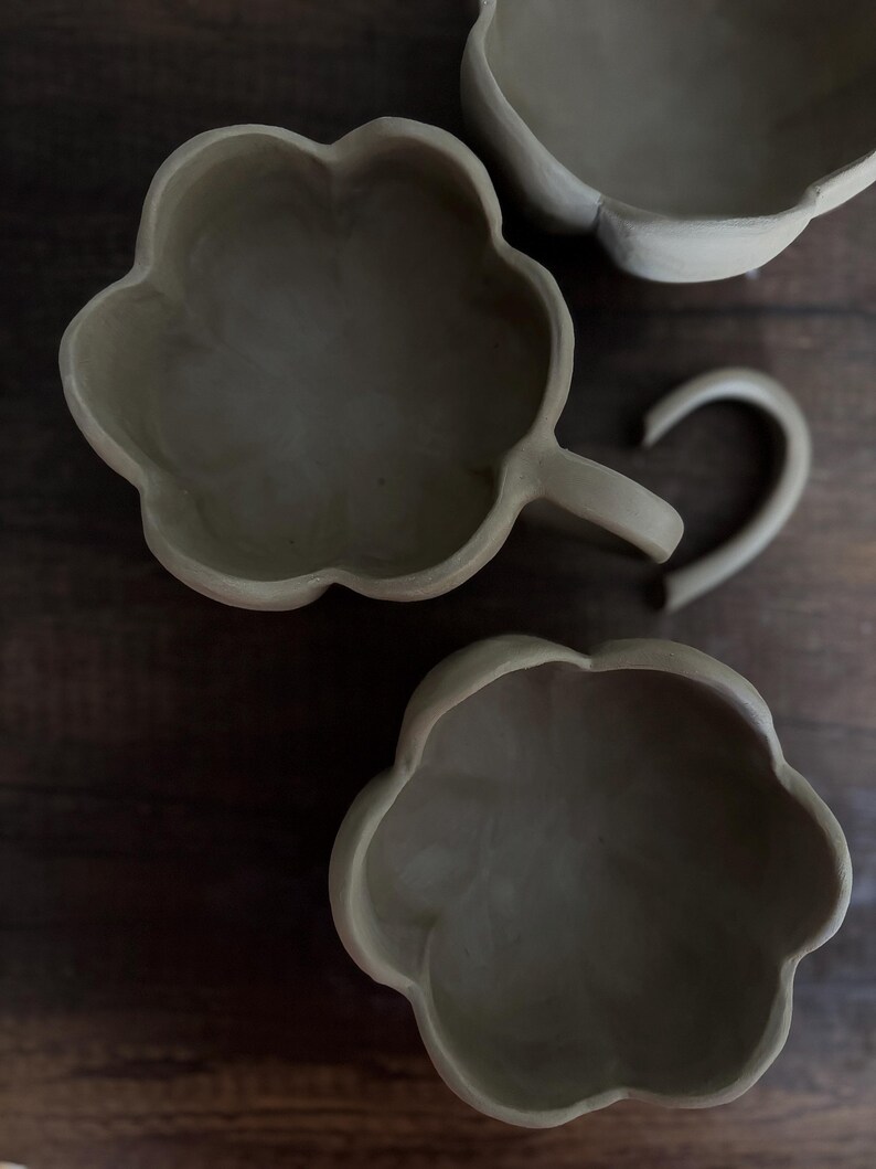 K&ouml;nnte beinhalten: Drei handgefertigte, unglasierte Keramikst&uuml;cke in Blumenform. Eines ist eine Tasse mit gebogenem Henkel, die anderen beiden sind Schalen. Die St&uuml;cke haben einen neutralen, erdigen Farbton, vor dunklem Holzgrund.