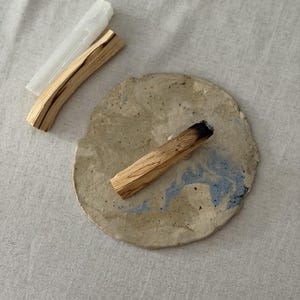 May include: A round, textured ceramic plate with beige and blue marbled patterns, featuring a charred wooden stick resting on top. Beside the plate are an additional unburnt wooden stick and a translucent, rectangular selenite crystal wand.