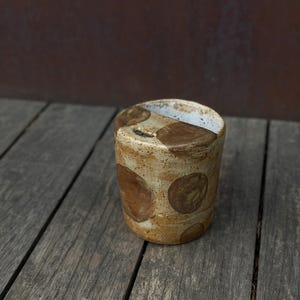 Op de afbeelding: Een cilindervormige keramische beker met een gespikkelde beige basis en grote, donkerbruine cirkelvormige patronen. De beker heeft een dikke, onregelmatige rand en een kleine, donkere inkeping aan de bovenkant, wat zorgt voor een ambachtelijke uitstraling.