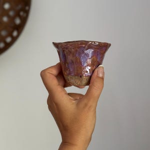 K&ouml;nnte beinhalten: Eine handgefertigte Keramiktasse mit ausgestelltem Rand. Die Tasse hat eine strukturierte, braune Au&szlig;enseite und eine gl&auml;nzende, violette Innenseite. Der Boden ist hellere, unglasierte Tonfarbe. Die Tasse wird in einer Hand gehalten.