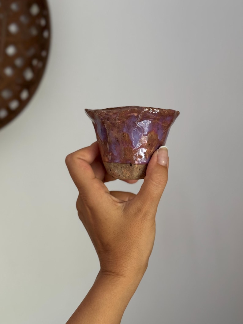 K&ouml;nnte beinhalten: Eine handgefertigte Keramiktasse mit ausgestelltem Rand. Die Tasse hat eine strukturierte, braune Au&szlig;enseite und eine gl&auml;nzende, violette Innenseite. Der Boden ist hellere, unglasierte Tonfarbe. Die Tasse wird in einer Hand gehalten.