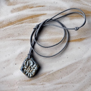 May include: A gray ceramic pendant with a gold floral design hangs on a gray cord necklace. The pendant is a square shape with rounded corners.