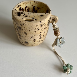 May include: A beige ceramic wall pocket with dark brown speckles, featuring a semi-circular opening at the top. A light-coloured cord hangs from the side, decorated with a brown wooden bead and two small, five-petaled ceramic flowers in blue and green.