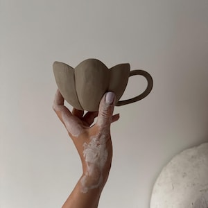 K&ouml;nnte beinhalten: Eine handgefertigte, hellbraune Keramiktasse mit einem einzigartigen tulpenf&ouml;rmigen Design. Die Tasse hat einen gebogenen Henkel und einen bl&uuml;tenblattartigen Rand. Die Tasse wird vor einem wei&szlig;en Hintergrund gehalten.