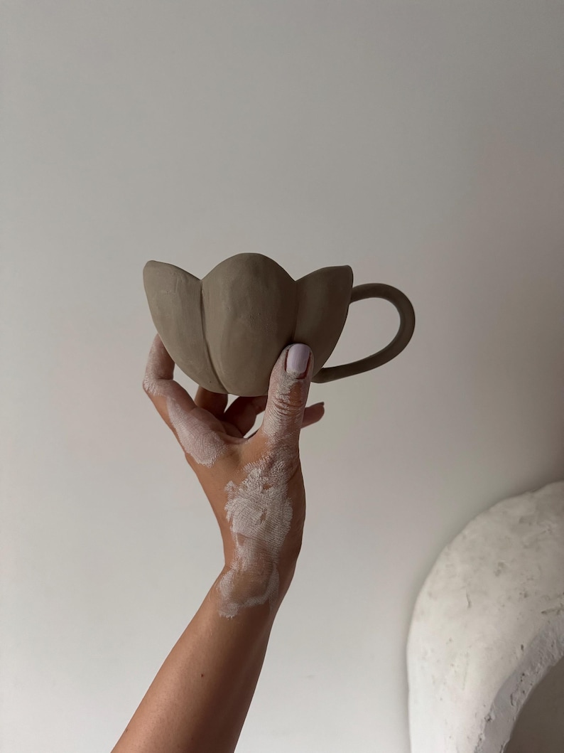 K&ouml;nnte beinhalten: Eine handgefertigte, hellbraune Keramiktasse mit einem einzigartigen tulpenf&ouml;rmigen Design. Die Tasse hat einen gebogenen Henkel und einen bl&uuml;tenblattartigen Rand. Die Tasse wird vor einem wei&szlig;en Hintergrund gehalten.