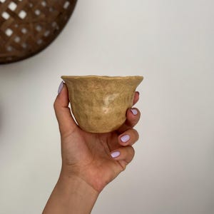 K&ouml;nnte beinhalten: Eine kleine, handgefertigte, beige Keramikschale mit strukturierter Oberfl&auml;che und gewelltem Rand. Die Schale wird in einer Hand mit hellviolettem Nagellack gehalten. Der Hintergrund ist eine schlichte wei&szlig;e Wand und ein geflochtener Korb.