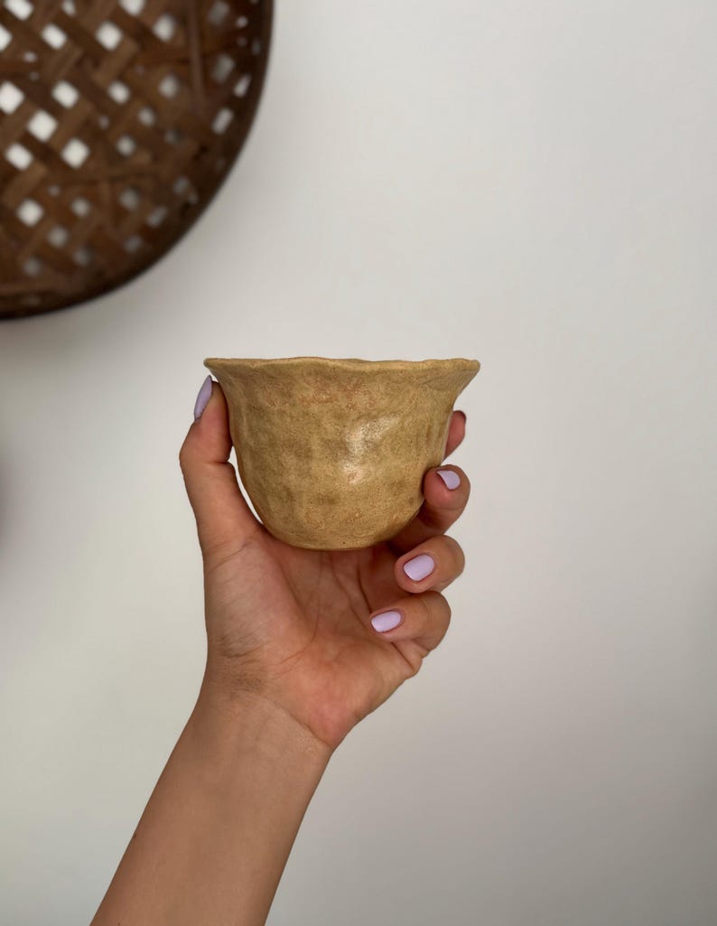 K&ouml;nnte beinhalten: Eine kleine, handgefertigte, beige Keramikschale mit strukturierter Oberfl&auml;che und gewelltem Rand. Die Schale wird in einer Hand mit hellviolettem Nagellack gehalten. Der Hintergrund ist eine schlichte wei&szlig;e Wand und ein geflochtener Korb.