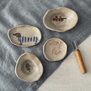 Op de afbeelding: Vier kleine, onregelmatige, gespikkelde keramische schaaltjes met handgeschilderde ontwerpen, waaronder een blauw gestreepte teckel, rode bessen, een rode abstracte vorm en een groene artisjok, vergezeld van een metalen vorkje met houten handvat.