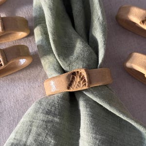 以下が含まれることがあります： セージグリーンのリネンナプキンが、ベージュのセラミック製ナプキンリングで留められています。リングには葉のようなテクスチャード加工が施されています。背景には他のリングも写っており、テーブルウェアのセットを連想させます。
