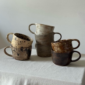 Könnte beinhalten: Eine Sammlung von sechs Keramikbechern mit Henkeln, die in drei Stapeln angeordnet sind. Die Becher weisen verschiedene Erdtöne wie Creme, Beige, Braun und Dunkelschokolade auf, mit gesprenkelten, gefleckten und einfarbigen Glasuroberflächen.