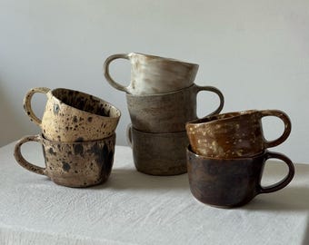 May include: A collection of handmade ceramic mugs in various earth tones. The mugs have a speckled, textured finish and curved handles. The colors range from light beige to dark brown, stacked on a white surface.