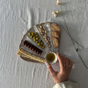 Könnte beinhalten: Eine muschelförmige Servierplatte mit einer Auswahl an Snacks: grüne Oliven, Pistazien, Sesam-Cracker und dunkle Schokolade. Eine kleine Schale mit Olivenöl und eine Hand mit Ringen sind ebenfalls zu sehen.