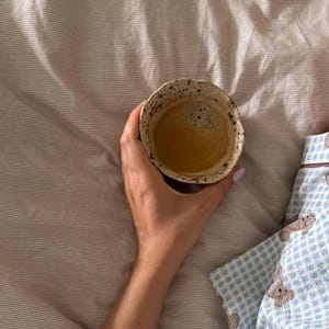 K&ouml;nnte beinhalten: Eine Hand h&auml;lt eine kleine, strukturierte Keramiktasse mit Kaffee. Der Kaffee hat eine schaumige Oberfl&auml;che. Die Tasse steht vor einem Hintergrund aus gestreifter Bettw&auml;sche und einem blau-wei&szlig; gemusterten Pyjama-Oberteil.