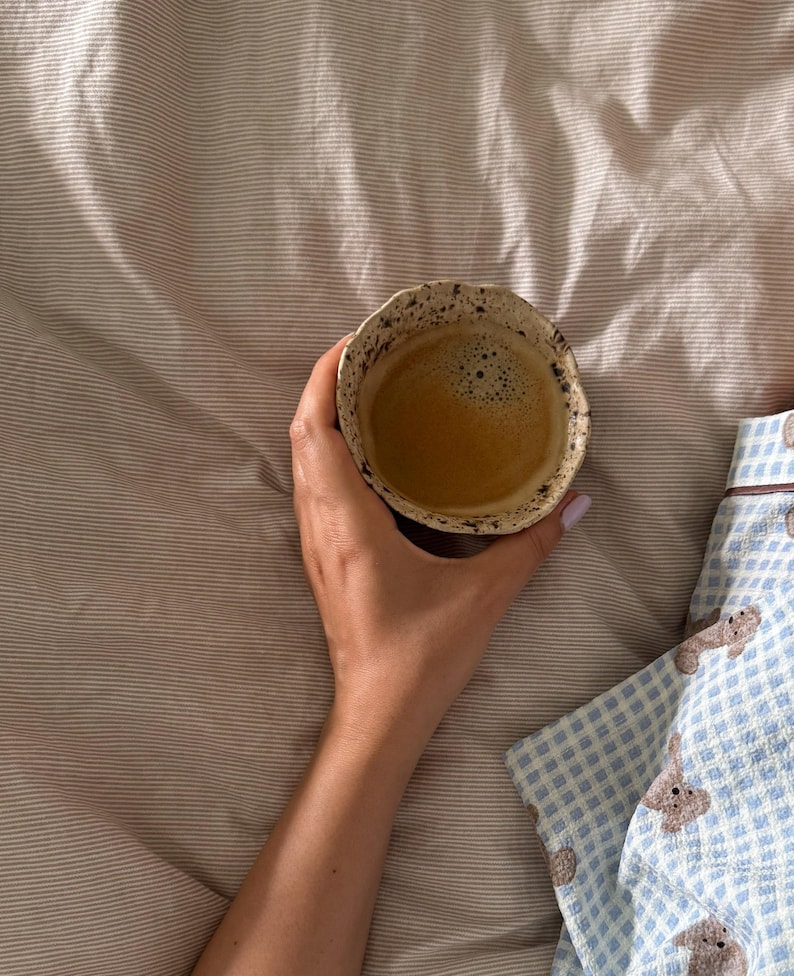 K&ouml;nnte beinhalten: Eine Hand h&auml;lt eine kleine, strukturierte Keramiktasse mit Kaffee. Der Kaffee hat eine schaumige Oberfl&auml;che. Die Tasse steht vor einem Hintergrund aus gestreifter Bettw&auml;sche und einem blau-wei&szlig; gemusterten Pyjama-Oberteil.