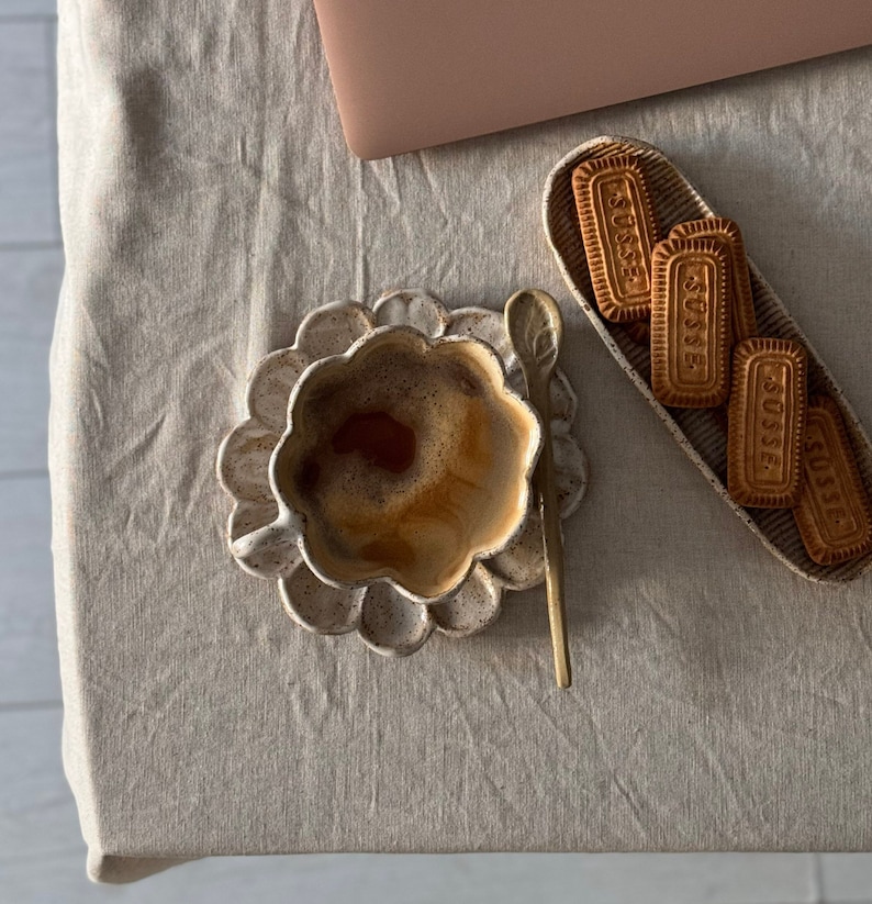 K&ouml;nnte beinhalten: Eine Kaffeetasse aus Keramik mit floralem Design, gef&uuml;llt mit Kaffee, steht auf einer passenden Untertasse. Ein kleiner goldener L&ouml;ffel liegt neben der Tasse. Ein rechteckiges Tablett enth&auml;lt mehrere Kekse mit dem Aufdruck "SUSSE".