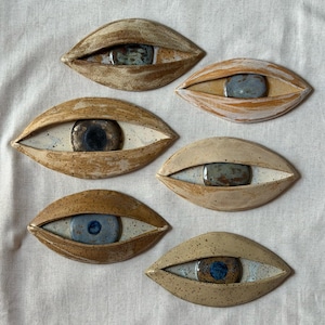 Op de afbeelding: Zes keramische oogvormige wanddecoraties in diverse tinten bruin, beige en zandkleur. Elk stuk heeft een centraal geglazuurde iris in blauwe, grijze of bruine tinten, omringd door een gespikkelde of gemarmerde kleistructuur als ooglid.
