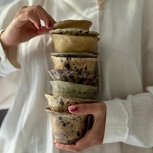 K&ouml;nnte beinhalten: Ein Stapel handgefertigter Keramikbecher in verschiedenen Braun-, Beige- und Gr&uuml;nt&ouml;nen. Die Becher haben ein gesprenkeltes Design und eine einzigartige, organische Form. Die Becher werden von den H&auml;nden einer Person gehalten.