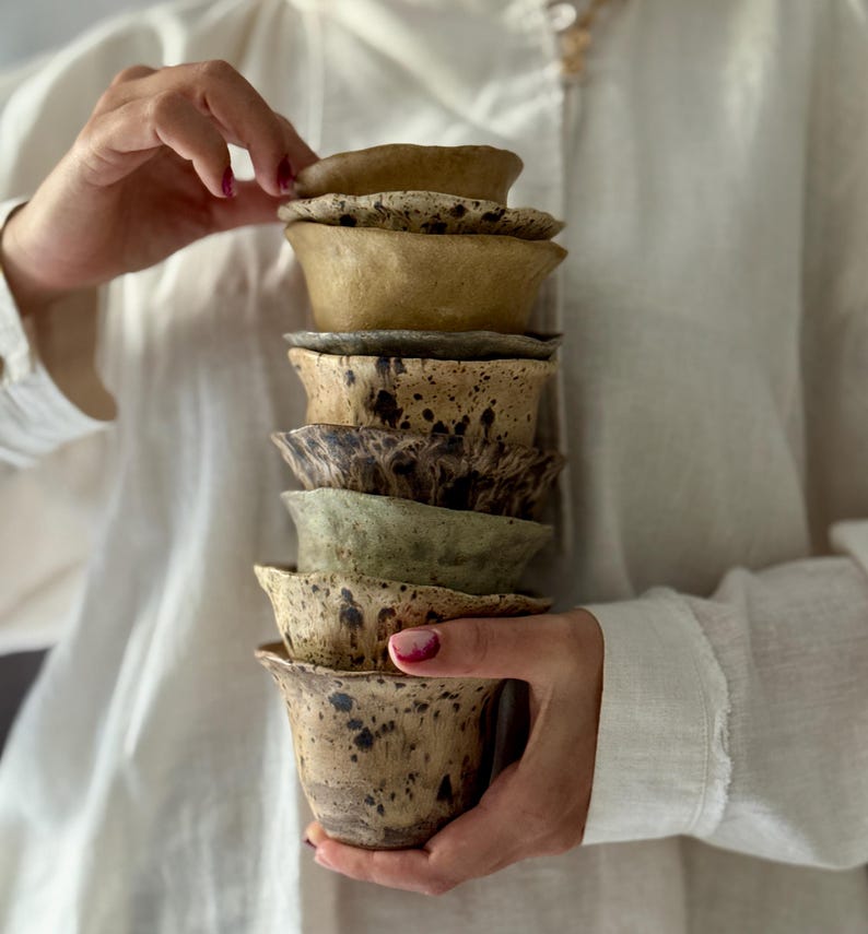 K&ouml;nnte beinhalten: Ein Stapel handgefertigter Keramikbecher in verschiedenen Braun-, Beige- und Gr&uuml;nt&ouml;nen. Die Becher haben ein gesprenkeltes Design und eine einzigartige, organische Form. Die Becher werden von den H&auml;nden einer Person gehalten.