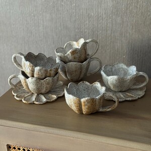K&ouml;nnte beinhalten: Ein Set aus sechs Keramik-Tassen und Untertassen. Die Tassen und Untertassen sind beige mit braunen Sprenkeln und einem gewellten Rand, der einer Blume &auml;hnelt. Die Tassen haben kleine Henkel. Das Set ist auf einer Holzoberfl&auml;che angeordnet.