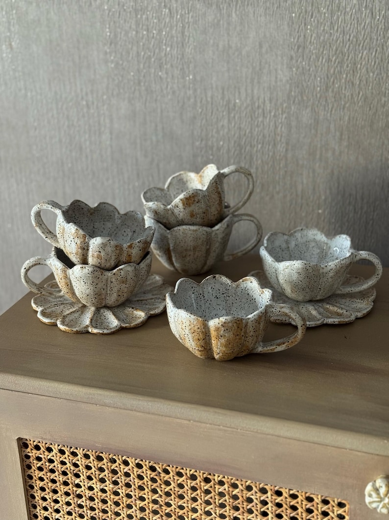K&ouml;nnte beinhalten: Ein Set aus sechs Keramik-Tassen und Untertassen. Die Tassen und Untertassen sind beige mit braunen Sprenkeln und einem gewellten Rand, der einer Blume &auml;hnelt. Die Tassen haben kleine Henkel. Das Set ist auf einer Holzoberfl&auml;che angeordnet.