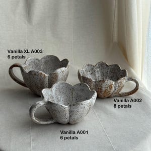 K&ouml;nnte beinhalten: Drei handgefertigte Keramikbecher mit einem Bl&uuml;tenblattdesign. Die Becher sind gesprenkelt wei&szlig; mit braunen Akzenten und haben einen Henkel. Der Text auf den Bechern lautet "Vanilla XL A003 6 petals", "Vanilla A002 8 petals" und "Vanilla A001 6 petals".