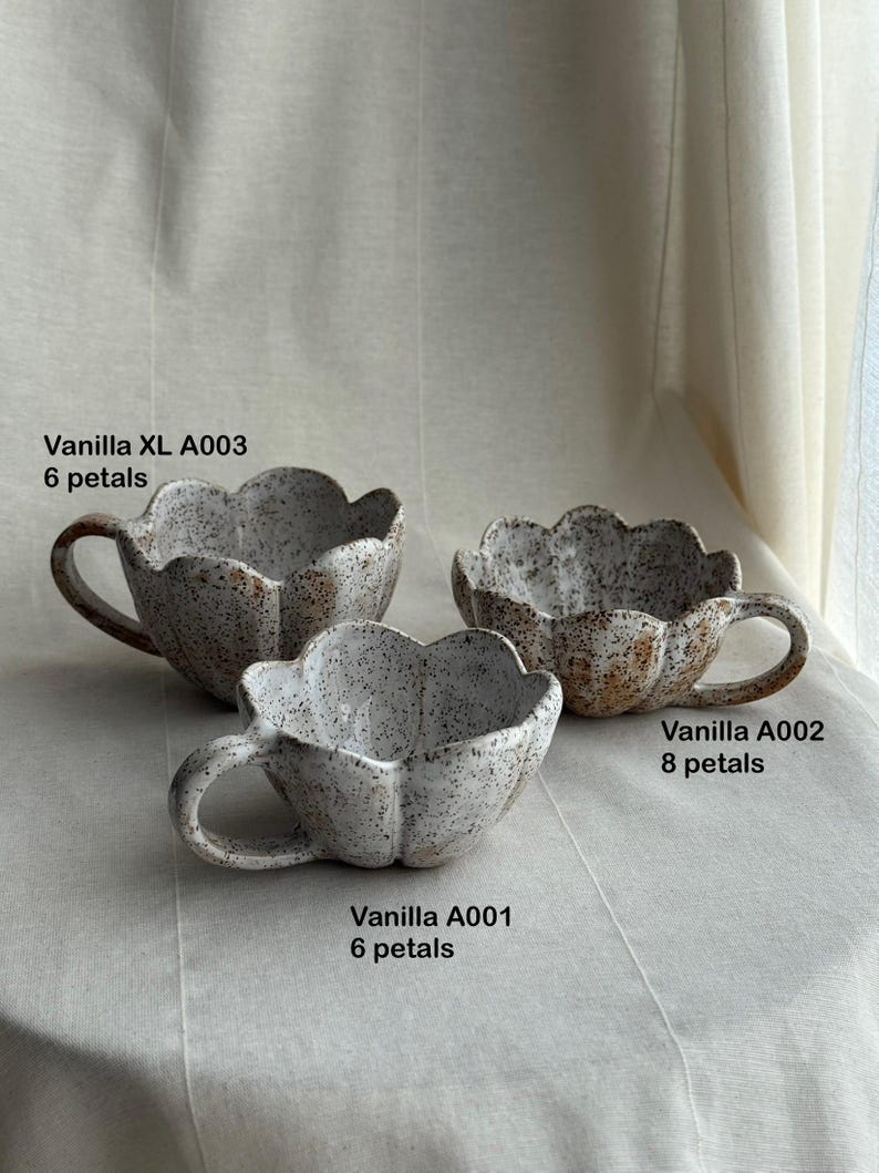 K&ouml;nnte beinhalten: Drei handgefertigte Keramikbecher mit einem Bl&uuml;tenblattdesign. Die Becher sind gesprenkelt wei&szlig; mit braunen Akzenten und haben einen Henkel. Der Text auf den Bechern lautet "Vanilla XL A003 6 petals", "Vanilla A002 8 petals" und "Vanilla A001 6 petals".