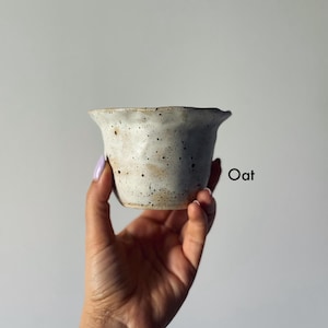 K&ouml;nnte beinhalten: Eine kleine, gesprenkelte Keramikschale in hellem Cremeton wird in einer Hand gehalten. Die Schale hat einen leicht unregelm&auml;&szlig;igen Rand und eine strukturierte Oberfl&auml;che. Das Wort "Oat" ist rechts auf der Schale schwarz aufgedruckt.
