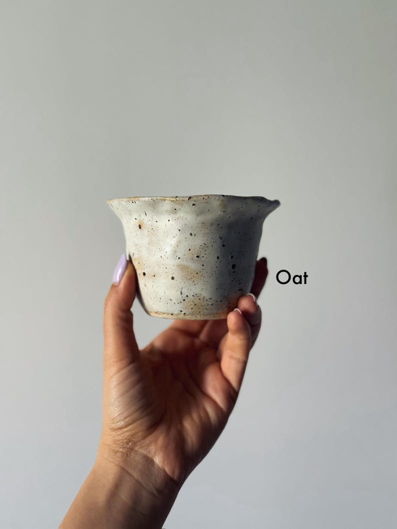 K&ouml;nnte beinhalten: Eine kleine, gesprenkelte Keramikschale in hellem Cremeton wird in einer Hand gehalten. Die Schale hat einen leicht unregelm&auml;&szlig;igen Rand und eine strukturierte Oberfl&auml;che. Das Wort "Oat" ist rechts auf der Schale schwarz aufgedruckt.