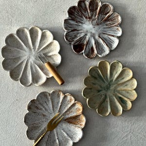 May include: Four ceramic plates with a flower petal design in cream, brown, and green. Two plates have small forks. The plates have a speckled finish and are arranged on a neutral-coloured surface.