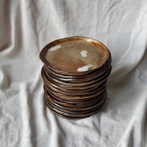 Może przedstawiać: Stos brązowych ceramicznych talerzy z białymi plamkami. Talerze są ułożone na białym lnianym materiale.