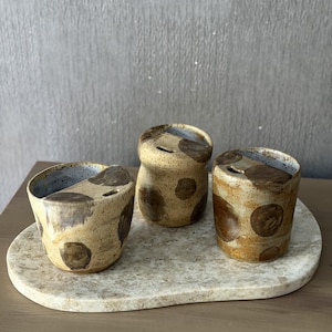 May include: Three small, cylindrical ceramic vessels with a speckled tan and brown finish featuring dark brown circular spots. Each vessel has a partially covered top with a narrow rectangular opening, revealing a light blue interior glaze.