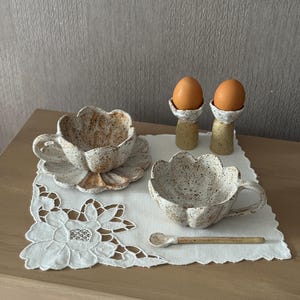 K&ouml;nnte beinhalten: Ein Keramik-Fr&uuml;hst&uuml;cks-Set mit einer bl&uuml;tenf&ouml;rmigen Tasse und Untertasse, einer passenden Sch&uuml;ssel und einem kleinen L&ouml;ffel. Zwei Eier liegen in gesprenkelten Eierbechern. Das Set befindet sich auf einer wei&szlig;en, floralen Tischdecke.