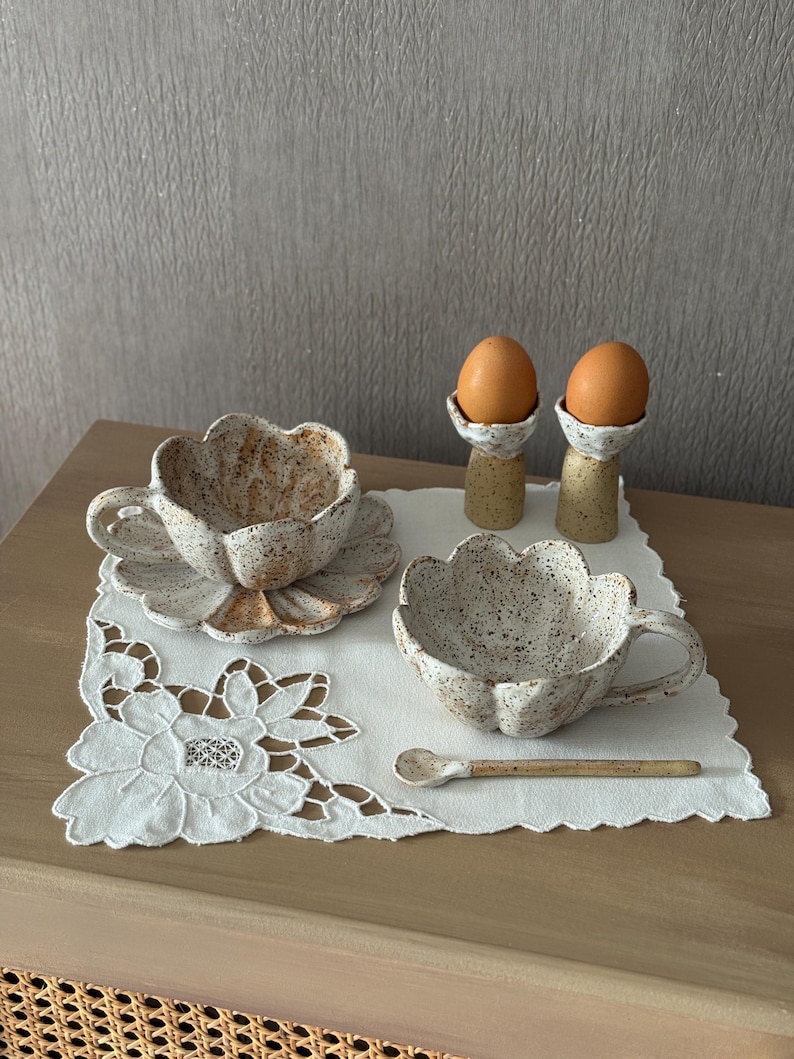 K&ouml;nnte beinhalten: Ein Keramik-Fr&uuml;hst&uuml;cks-Set mit einer bl&uuml;tenf&ouml;rmigen Tasse und Untertasse, einer passenden Sch&uuml;ssel und einem kleinen L&ouml;ffel. Zwei Eier liegen in gesprenkelten Eierbechern. Das Set befindet sich auf einer wei&szlig;en, floralen Tischdecke.