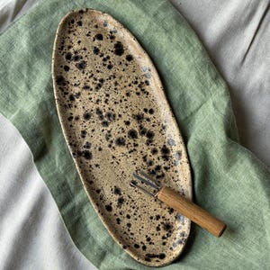 May include: An oval ceramic serving platter with a speckled tan and black pattern. A small fork with a wooden handle is on the platter. The platter is on a green and beige linen surface.