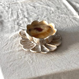 K&ouml;nnte beinhalten: Ein Keramik-Kaffeebecher und Untertassen-Set. Der Becher und die Untertasse sind wie eine Blume mit einem gewellten Rand geformt. Der Becher ist mit Kaffee gef&uuml;llt. Das Set ist cremefarben mit braunen Sprenkeln.