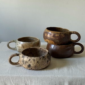 Könnte beinhalten: Vier handgefertigte Keramikbecher mit abgerundeten Körpern und angebrachten Henkeln. Die Becher sind in Braun-, Beige- und Beigetönen gehalten und haben gesprenkelte und glasierte Oberflächen. Sie sind auf einer neutralfarbenen Oberfläche angeordnet.