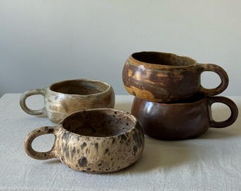 Peut inclure: Quatre tasses en céramique faites à la main, avec des corps arrondis et des anses attachées. Les tasses sont dans des tons de marron, beige et tan, avec des finitions mouchetées et vernissées. Elles sont disposées sur une surface de couleur neutre.