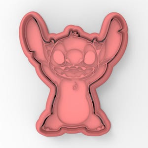 May include: Pink plastic cookie cutter shaped like Stitch from the Disney movie Lilo & Stitch. Stitch is standing with his arms raised and has a mischievous expression on his face.