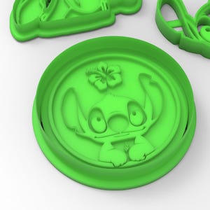 May include: Green plastic cookie cutter featuring a cartoon character with large eyes and a flower. The character is Stitch from the Disney movie Lilo & Stitch.