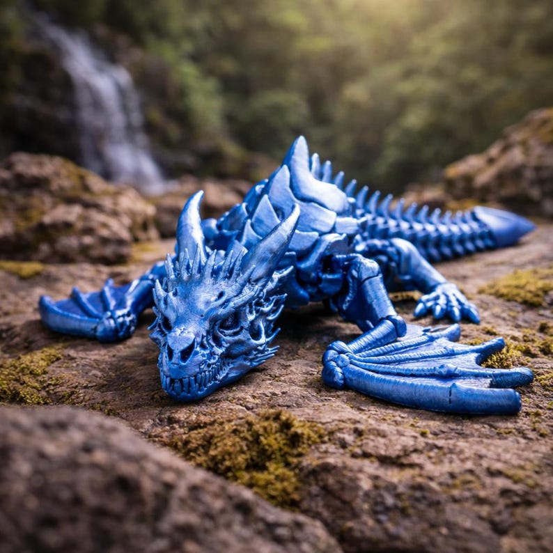 May include: A detailed, blue dragon figurine with textured scales, horns, and wings. The fantasy creature is posed on a rocky surface, with a waterfall and greenery in the background. The dragon is a vibrant shade of blue.