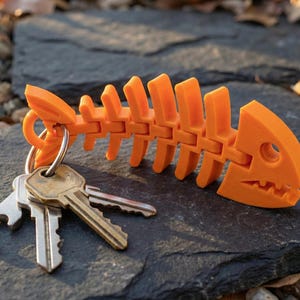 May include: An orange fish skeleton keychain with a silver keyring and several keys. The fish skeleton is made of plastic and has a detailed design. The keychain is resting on a dark gray stone surface.