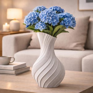 Puede incluir: Un jarrón de cerámica blanca con un diseño en espiral contiene un ramo de flores de hortensia azules. El jarrón está sobre una mesa de madera, con un sofá de tonos neutros en el fondo. Las flores tienen hojas verdes.