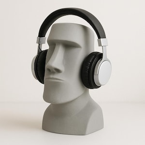 Puede incluir: Una estatua gris de Moai con auriculares negros y plateados. Los auriculares tienen auriculares negros y una diadema negra. La estatua es un busto de una cabeza grande con una expresión severa.