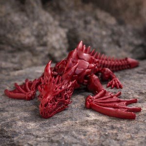 May include: A red dragon figurine with detailed scales, horns, and wings. The fantasy creature is posed on a textured, gray surface. The dragon's design includes a long tail and sharp claws.