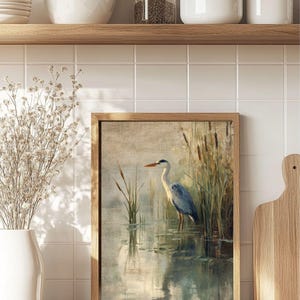 May include: A framed painting of a grey heron standing in a pond with reeds in the background. The heron is facing to the right and has its head turned to the left. The water is a light blue color and the reeds are a light green color.