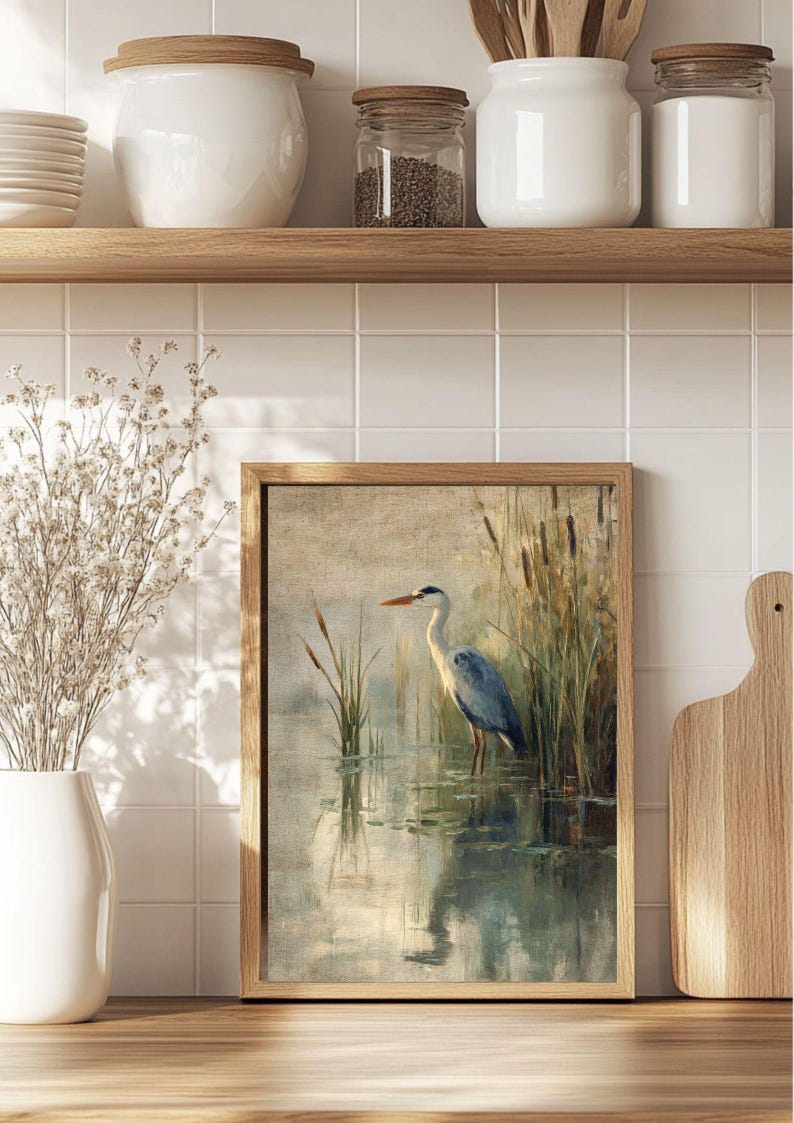 May include: A framed painting of a grey heron standing in a pond with reeds in the background. The heron is facing to the right and has its head turned to the left. The water is a light blue color and the reeds are a light green color.