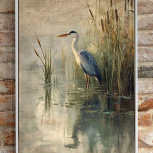 May include: A framed painting of a grey heron standing in a pond with reeds in the background. The heron is facing to the left and has its head turned to the right. The water is a light blue-green color and the reeds are a light brown color.