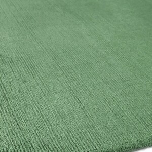 Pu&ograve; includere: Primo piano di un tappeto verde con una superficie strutturata. Il tappeto ha un colore verde chiaro e un motivo nell'angolo superiore con forme nere e bianche. La trama del tappeto &egrave; visibile.