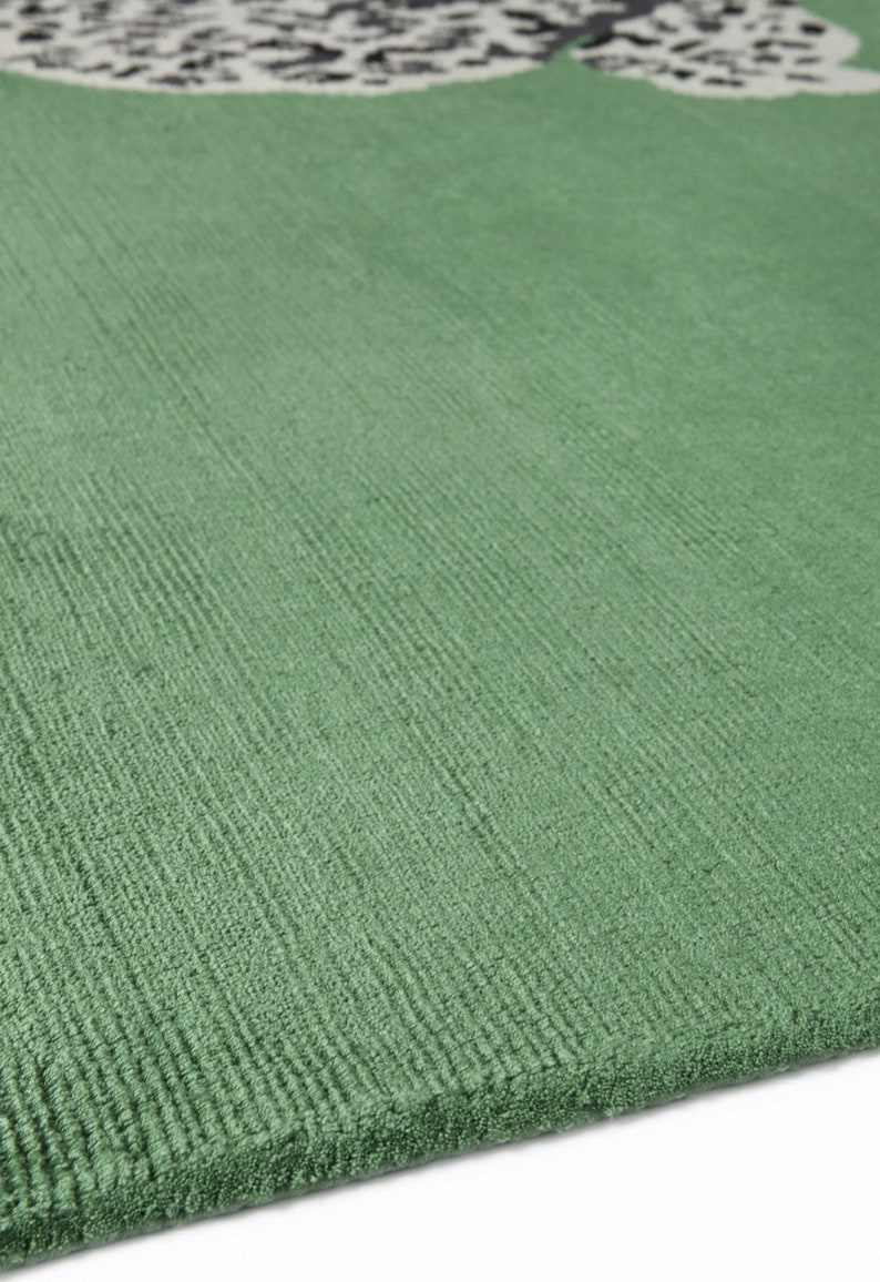 Pu&ograve; includere: Primo piano di un tappeto verde con una superficie strutturata. Il tappeto ha un colore verde chiaro e un motivo nell'angolo superiore con forme nere e bianche. La trama del tappeto &egrave; visibile.