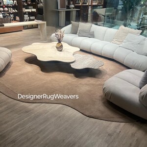 Op de afbeelding: Een groot, lichtbruin, pluche tapijt met een unieke, organische vorm. Het tapijt bevindt zich in een woonkamer met een witte bank, twee salontafels en een vaas met gedroogde bloemen. De tekst "DesignerRugWeavers" is zichtbaar op het tapijt.